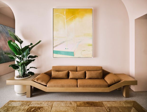 A warm, refined living room with a large low-profile brown upholstered sofa, abundant indoor greenery, a pale blush curved architectural wall, abstract artwork, and a rustic patterned area rug. The room has a sculptural mid-century-inspired feel with soft natural tones and rounded openings.