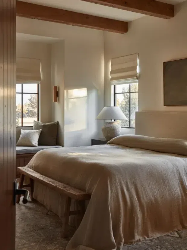 A warm, softly lit bedroom with a large upholstered bed dressed in layered beige linen bedding. The room features exposed timber ceiling beams, white plaster walls, a built-in window seat with cushions, Roman blinds, a textured bedside lamp, a simple artwork panel, and a rustic timber bench at the foot of the bed. A patterned neutral rug adds texture underfoot.