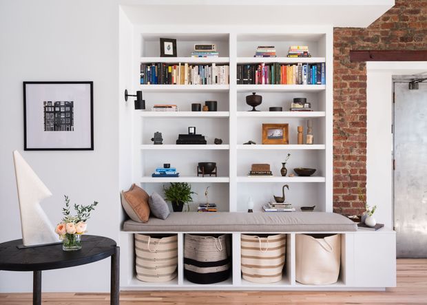 Bright, carefully styled living room with a large built-in white shelving and cabinetry unit filled with books and decorative objects. A cushioned window seat or built-in bench runs across the lower section, with woven storage baskets beneath. The room includes a round dark table, floor lamp, framed wall art, potted plant, and an exposed red-brick wall with a metal-framed door, creating a refined contemporary interior with industrial accents.