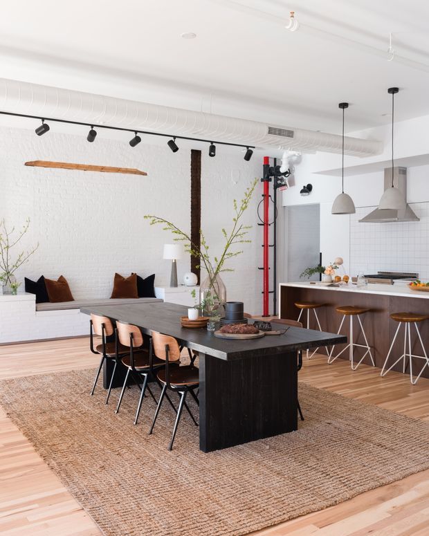 Bright open-plan loft-style interior combining a dining area and kitchen. A long dark dining table with multiple chairs sits on a large natural-fibre rug, while the kitchen features a dark island with bar stools, white tiled walls, pendant lights, and stainless-steel appliances. White painted brick walls, exposed ductwork, track lighting, timber flooring, and a red climbing-style feature add an industrial character softened by greenery and warm wood tones.