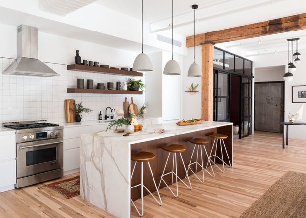 Bright, spacious open-plan kitchen with a large white marble-look island and peninsula seating, stainless steel range and hood, white tiled splashback, open timber shelves, and multiple pendant lights. Warm timber flooring and exposed reclaimed timber beams contrast with the white cabinetry and walls. A black-framed glazed partition and adjoining living or dining area are visible beyond the kitchen.
