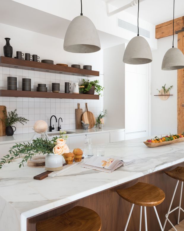 Bright, refined contemporary kitchen with a large white marble island and timber cabinetry, open shelving against a white square-tile backsplash, matte black tapware, and three sculptural concrete pendant lights. Warm timber wall cladding and stools add contrast, while the island is styled with flowers, fruit, and open books.