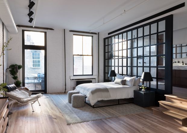 A bright industrial-style bedroom with a low platform bed, white bedding, black bedside tables and lamps, and a soft area rug over wide-plank timber flooring. Large black-framed steel-look glazing forms a partition behind the bed, separating the bedroom from an adjacent kitchen or living area. White-painted brick walls, exposed black conduit, track lighting, tall windows, and a timber stair add architectural character.
