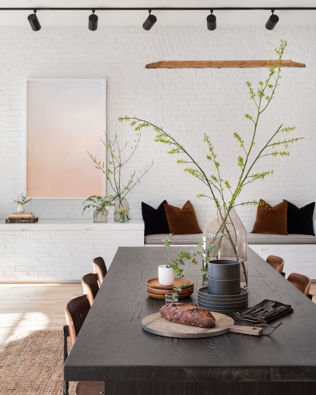 A bright contemporary dining room with a long dark timber dining table set for a meal, surrounded by warm brown leather dining chairs. A white-painted exposed brick wall forms the backdrop, with a built-in bench seat styled with black and rust cushions. Track lighting, a floating timber shelf, large pastel artwork, and tall leafy branches add contrast and texture.