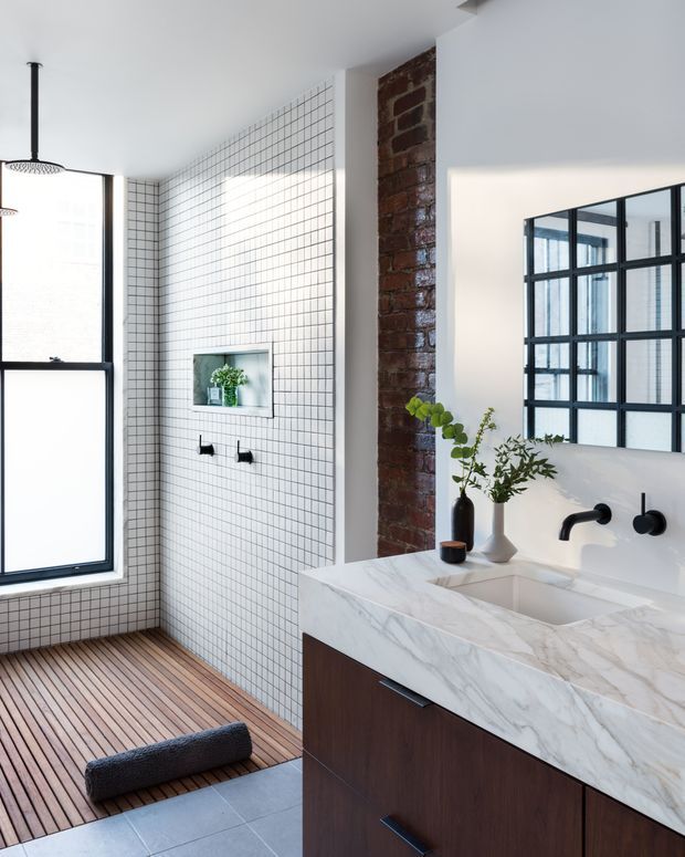 Bright contemporary bathroom featuring a walk-in shower lined with small white square tiles, a timber slatted floor, exposed red brick, and a dark wood vanity with a thick white marble benchtop. Black-framed glazing and a matching black-framed mirror add an industrial accent, while potted greenery provides a natural touch.