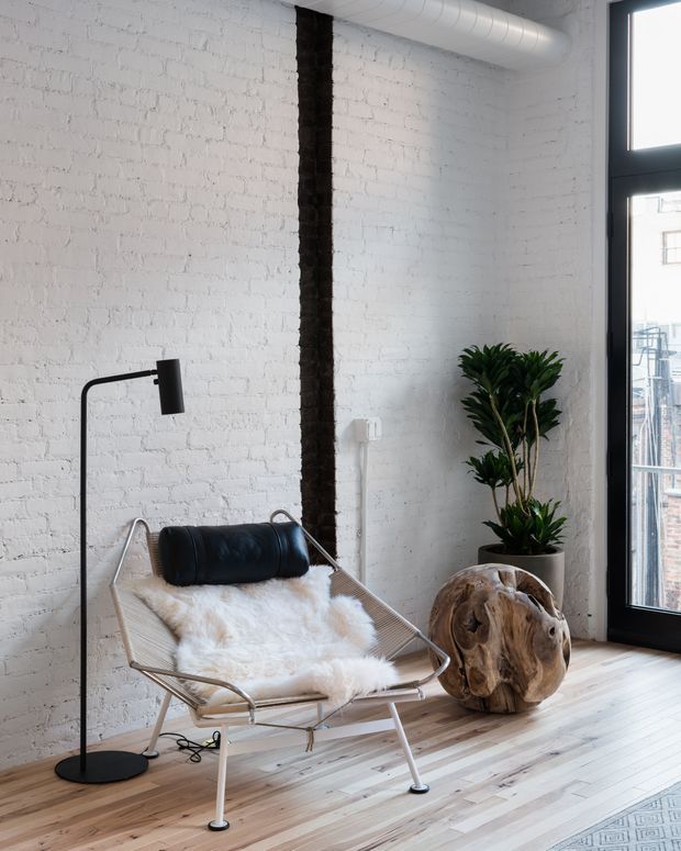 Bright loft-style interior corner featuring a black leather and metal sling chair draped with a white sheepskin, a sculptural black floor lamp, a potted indoor tree, and a rustic carved wood accent on pale timber flooring. White-painted exposed brick walls, black-framed floor-to-ceiling glazing, and an exposed ceiling duct create an industrial aesthetic.