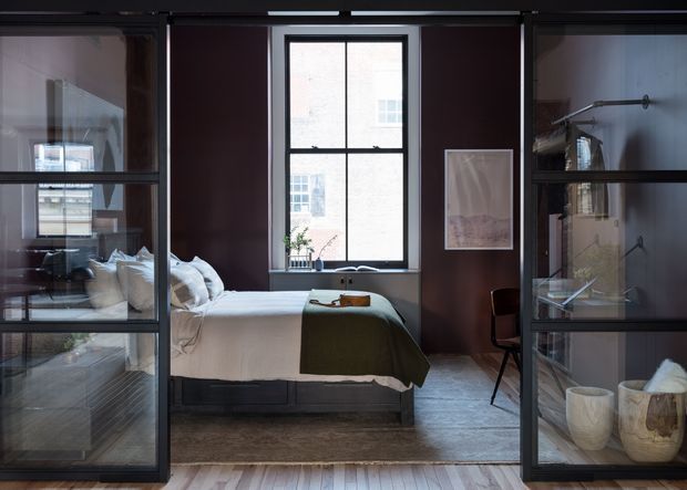 A compact, refined bedroom viewed through dark-framed glass-and-metal partition doors. The room features a neatly made bed with white bedding and an olive-green throw, deep brown walls, a central black-framed window, a simple chair, artwork, and open shelving displaying neutral-toned objects. Timber flooring and layered glazing create a sophisticated, intimate atmosphere.