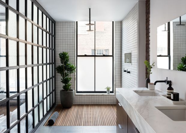 Bright contemporary bathroom with a long double vanity, white marble-look benchtop, wall-mounted black fixtures, white small-format grid tiles, and a large industrial-style black-framed glazed partition. A timber slatted floor area, indoor plants, exposed brick column, and generous windows add warmth and texture.