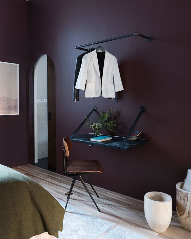 A moody bedroom corner with deep burgundy walls, a floating black wall-mounted desk, a mid-century style wooden chair, and a wall-mounted clothing rail holding jackets. An arched doorway opens to a tiled bathroom or adjoining space. Timber flooring, a pale patterned rug, framed artwork, plants, and ceramic vessels complete the scene.