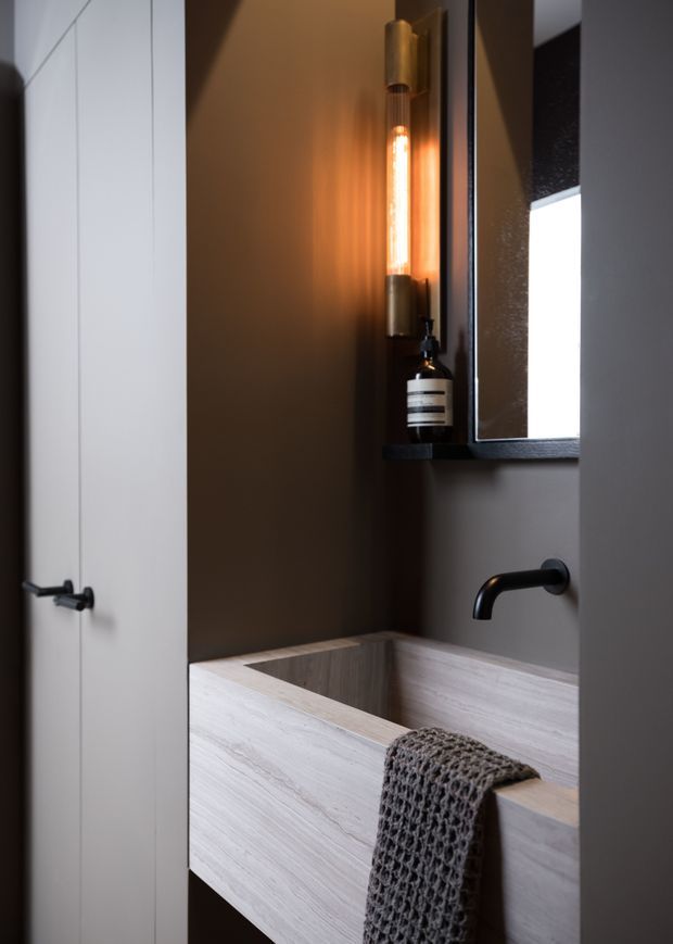 A compact, moody bathroom with a wall-mounted light timber vanity featuring an integrated rectangular basin, matte black wall-mounted tapware, dark painted walls, a black-framed mirror, and a warm vertical feature light. A textured charcoal hand towel hangs over the vanity.