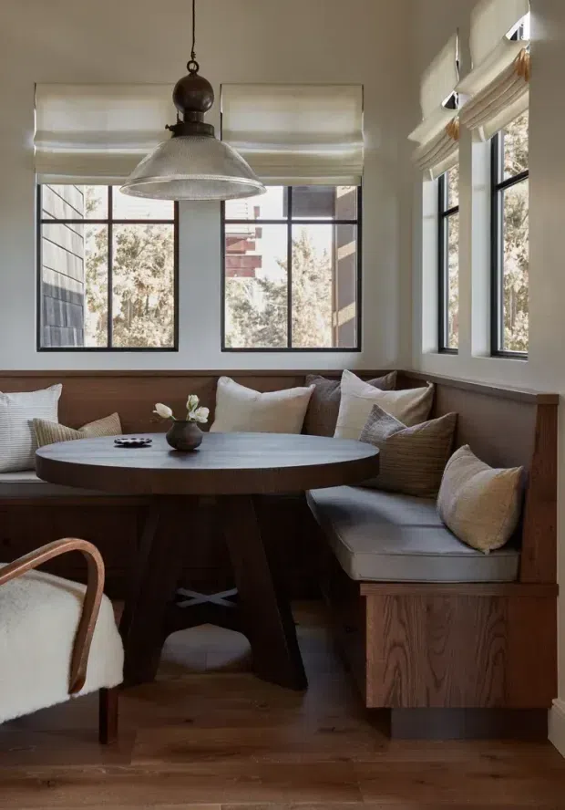 A warm, compact dining nook with a built-in wood banquette wrapping around a round dark wood table. Neutral cushions, a sculptural pendant light, framed windows with Roman blinds, white walls, and timber flooring create a refined, inviting interior.