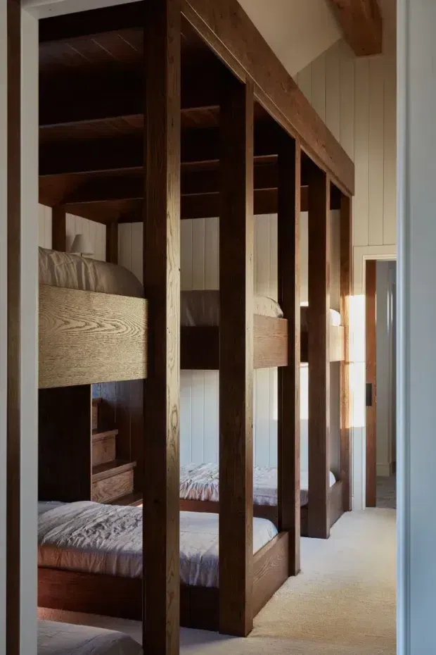 A compact bunk bedroom viewed through a doorway, featuring multiple built-in timber bunk beds arranged along the walls. Dark stained exposed timber posts and beams create a strong structural framework, while light vertical wall panelling, pale carpet, and neutral bedding soften the room. A doorway and bright natural light are visible at the rear.