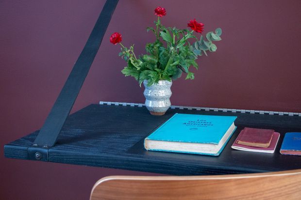 Close-up of a dark timber wall-mounted shelf or desk against a deep burgundy wall. The surface holds a textured ceramic vase with red flowers and greenery, several books, and a small notebook. A curved timber chair back is visible in the foreground, suggesting a compact study or workspace. The image has a moody, refined mid-century-inspired character with strong burgundy, dark wood, and turquoise accents.