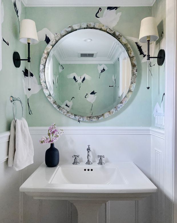 A compact, elegantly styled traditional powder room with a white pedestal vanity and integrated rectangular basin, polished chrome three-piece tapware, pale sage-green walls, white wainscoting and moulded trim. A large round mirror with a textured stone-like frame dominates the wall, flanked by black wall-mounted sconces with cream shades. Crane-patterned wallpaper or mural detailing adds a playful decorative accent, while a towel ring and dark vase with flowers complete the room.