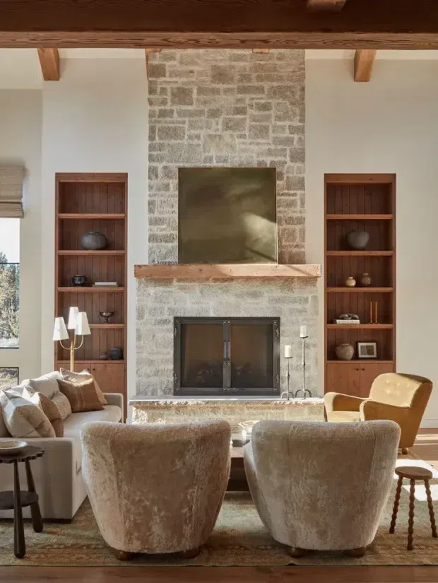 Warm, refined living room centred on a large natural-stone fireplace with timber mantel and exposed timber ceiling beams. Built-in dark timber shelving flanks the fireplace, while upholstered lounge chairs, a sofa, side tables and layered neutral textiles create a comfortable sitting area. Natural light enters from the windows at left.
