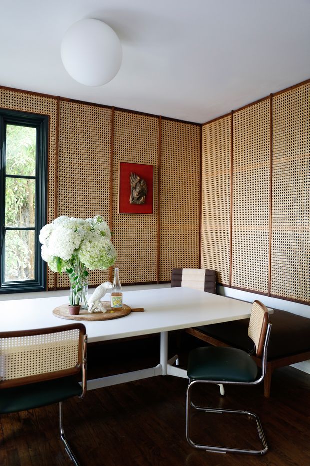 A refined dining nook with a white built-in corner banquette and rectangular dining table, surrounded by warm woven cane/rattan wall panelling in dark timber frames. Matching cane-and-chrome cantilever chairs with dark green seats sit around the table. A black-framed window, white globe ceiling light, artwork, and a floral arrangement complete the room.