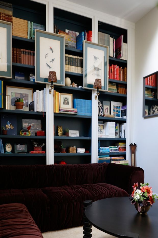 A richly styled living room with dark built-in bookshelves spanning the wall, filled with books, framed artwork, photographs, and decorative objects. A deep burgundy velvet sectional sofa surrounds a black rounded coffee table topped with a colourful floral arrangement. White wall and shelving trim, framed bird illustrations, and a wall mirror complete the eclectic, book-lined interior.