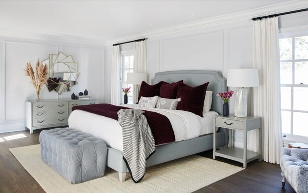 Bright, refined bedroom with a pale upholstered bed, white bedding, deep burgundy accent pillows and throw, a tufted bench, matching light bedside tables, decorative wall panelling, full-length curtains, and dark timber flooring. The room has abundant natural light and a polished, comfortable appearance.