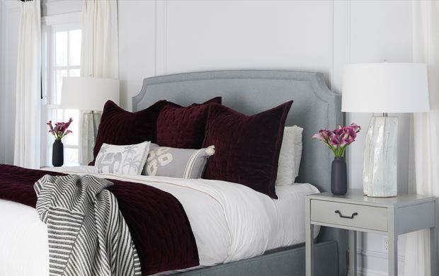 Bright, refined bedroom featuring a pale grey upholstered bed with a curved headboard, crisp white bedding, deep burgundy velvet pillows and throw, patterned accent cushions, white bedside tables, ceramic lamps, floral arrangements, and tall curtained windows. The room has a balanced, elegant contemporary-traditional feel.
