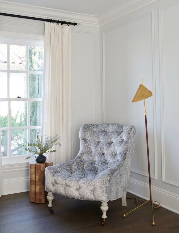 A bright, refined sitting corner featuring a tufted grey upholstered armchair on turned legs, a sculptural wood side table with a small vase, and a slender brass-and-wood floor lamp. The room has pale grey decorative wall panelling, white crown moulding, a tall multi-pane window with cream curtains, and dark timber flooring.