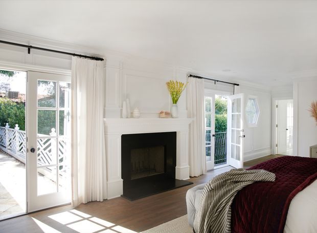 Bright, elegant bedroom with white paneled walls, a traditional decorative fireplace, tall French doors opening to a balcony or deck, and large windows dressed with white curtains. A bed with a deep burgundy coverlet and layered throws occupies the foreground, while natural timber flooring and abundant daylight create an airy, refined atmosphere.