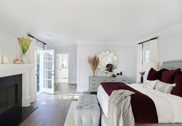 Bright, elegant bedroom with a large upholstered bed dressed in white and deep burgundy bedding, a tufted bench, white paneled walls and ceiling, dark timber flooring, and a traditional fireplace. Tall French doors and windows with sheer curtains provide abundant natural light. A white dresser and geometric wall mirror complete the refined interior.