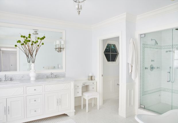 Bright, elegant white bathroom with a large double vanity, mirrored wall, decorative chandelier-style lighting, freestanding shower enclosure, and a small white dressing stool. The room features detailed cabinetry, wall moulding, a patterned tiled floor, and a traditional glass-panel door.