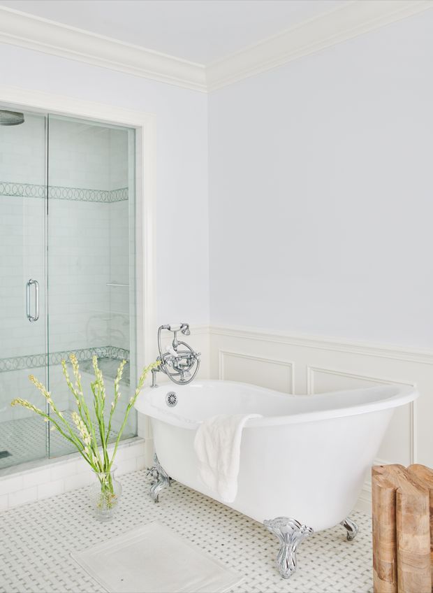 Bright, elegant bathroom featuring a freestanding white clawfoot bathtub, glass-enclosed shower, pale mosaic floor tiles, white tiled shower walls with a decorative border, chrome fixtures, and traditional wall moulding with framed panel details. A small wooden stool and a vase of greenery add warmth.