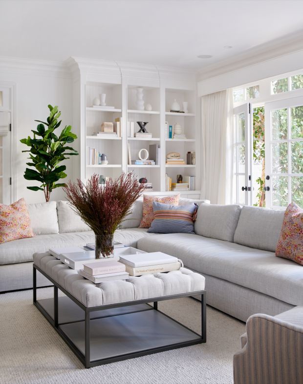 Bright, refined living room with a large light-grey sectional sofa arranged around a tufted ottoman coffee table with a dark metal frame and lower shelf. White built-in shelving displays books and decorative objects, while tall French doors and windows bring in abundant natural light. Neutral furnishings are accented by patterned and striped cushions, a large indoor plant, and a dried botanical arrangement.