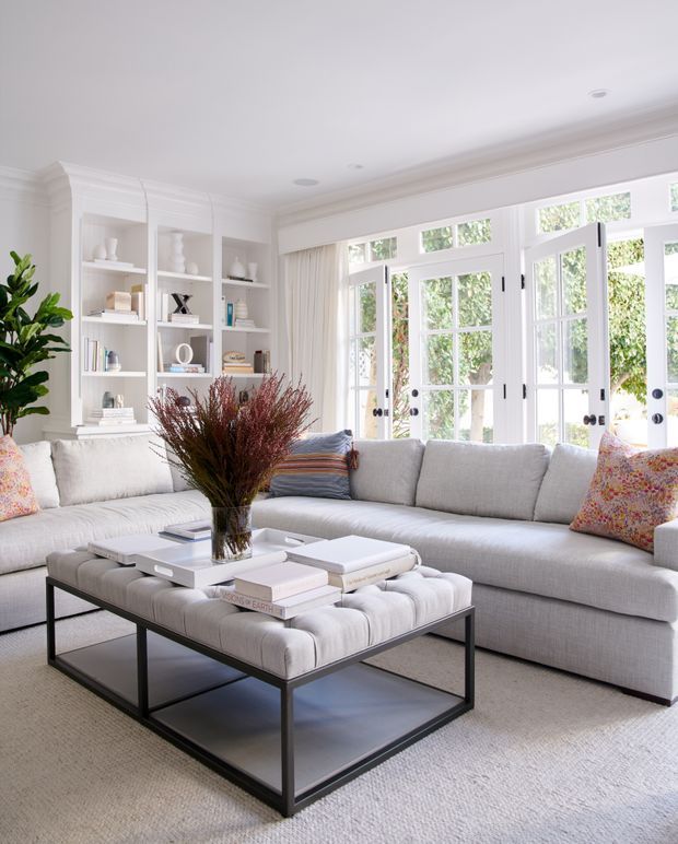 Bright, refined living room with a large light-grey sectional sofa, tufted square ottoman coffee table, white built-in display shelving, extensive white-framed French doors and windows, and layered neutral furnishings with colourful patterned cushions. The room has abundant natural light, high decorative ceiling moulding, and a calm contemporary-classic feel.