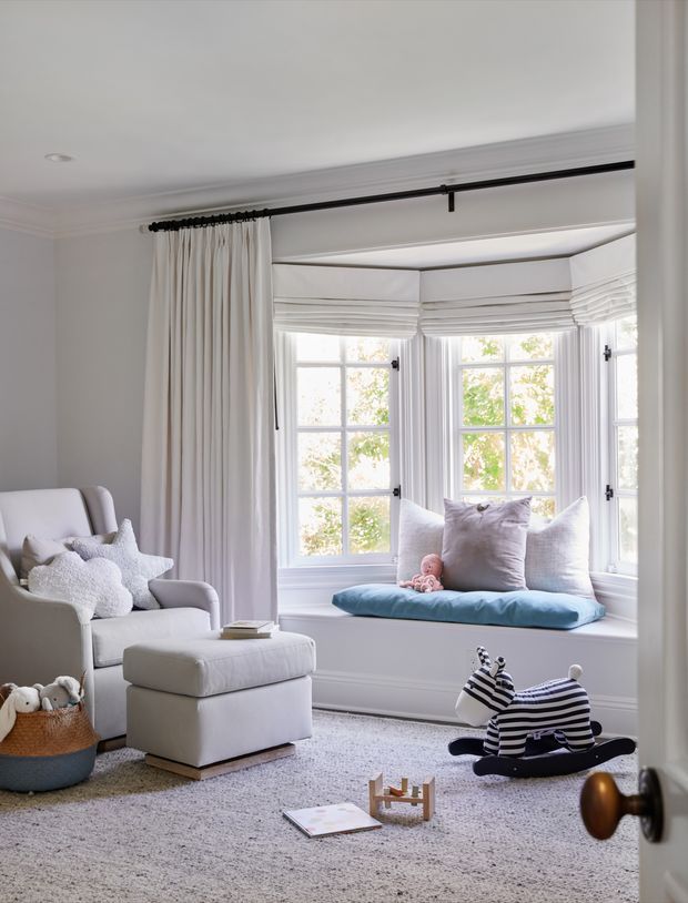 A bright, cozy nursery or child's room with a large white upholstered armchair and ottoman, a deep bay-window seat with cushions, and full-height white curtains on a dark curtain rod. The room features soft neutral finishes, plush carpeting, children's toys, and a small infant resting on the window seat. Traditional multi-pane windows and detailed white trim give the space a refined, classic character.