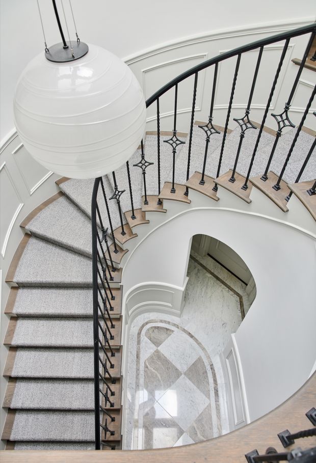A grand curved stairway viewed from above, featuring grey carpeted steps, pale timber stair nosings, ornate black metal balustrades, white walls with decorative panel moulding, and a large spherical pendant light. A marble-patterned tiled floor is visible in the lower level through the stairwell opening.