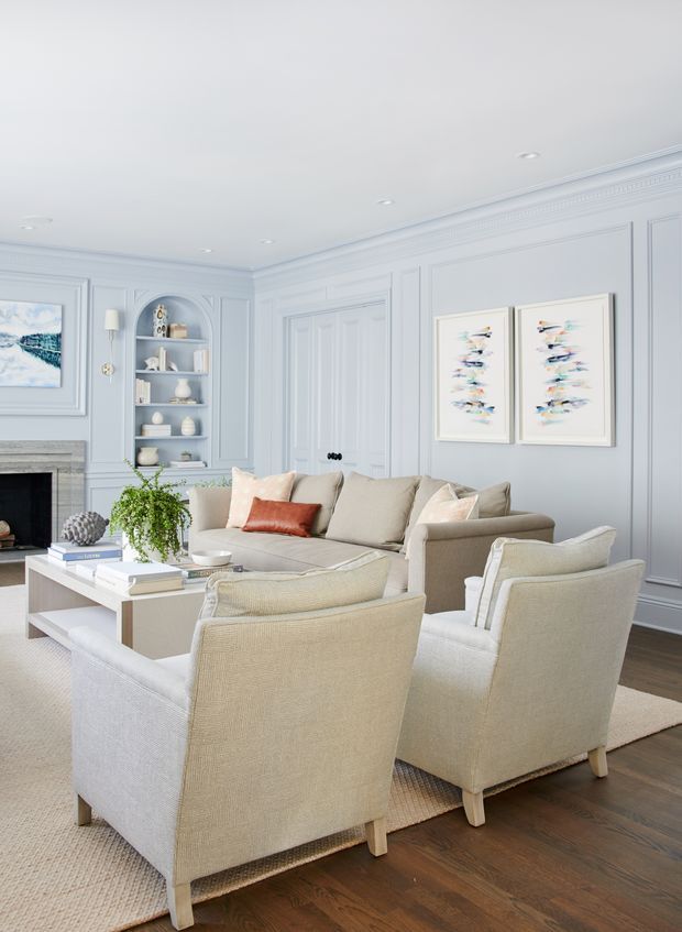 Bright, refined living room with pale blue-gray paneled walls, ornate crown moulding, a white cofferless ceiling with recessed lighting, built-in display shelving, a classic fireplace, and a closed paneled door. The room is furnished with a neutral upholstered sofa and armchairs arranged around a white coffee table, with layered rugs, light artwork, and a small indoor plant. Dark timber flooring provides contrast to the light, calm palette.