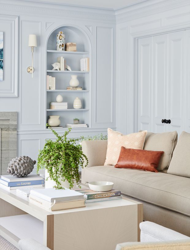 A bright, refined living room with pale blue-grey decorative wall panelling, ornate moulding, a built-in arched display niche, and paneled double doors. A neutral upholstered sofa with peach and rust cushions faces a light timber coffee table styled with books, a bowl, and a trailing plant. The room has a soft, elegant traditional character with layered architectural detailing and a calm, airy palette.