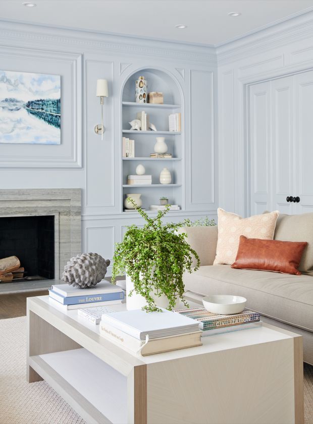 Bright, refined living room with pale blue-grey decorative wall panelling, elaborate crown moulding, built-in arched shelving, a marble or stone fireplace, and a light neutral sofa. A layered square coffee table with books, a potted trailing plant, ceramics and cushions adds a soft, elegant residential feel.