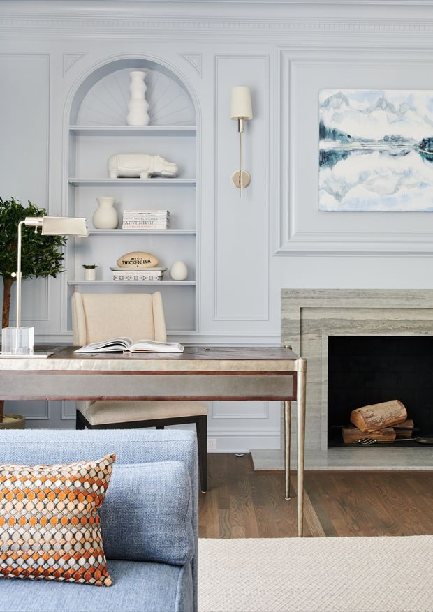 A refined living room and home office corner with pale blue-gray decorative wall paneling, built-in arched shelving, a writing desk, upholstered chair, blue sofa, patterned cushion, fireplace, and framed watercolor artwork. The room combines classic architectural detailing with elegant contemporary furnishings and a soft, cool-toned palette.