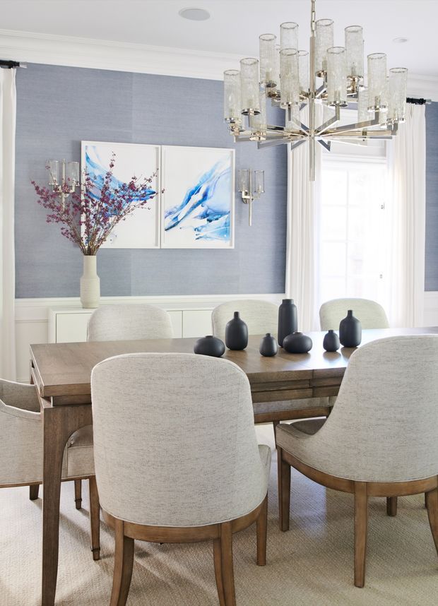 A bright, refined dining room with a large rectangular timber dining table surrounded by upholstered dining chairs. The room features blue textured wallcovering above white wall panelling, white curtains, abstract blue artwork, a substantial contemporary glass-and-metal chandelier, and dark decorative ceramic vessels arranged on the table.