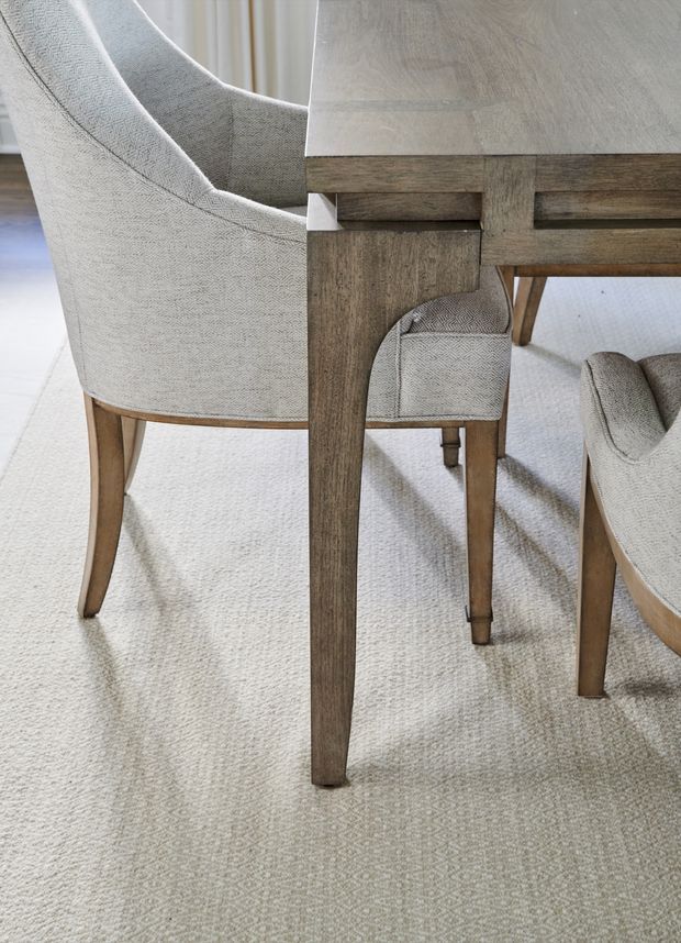 Close-up interior view of a dining setting, showing a rustic timber dining table with visible joinery, upholstered dining chairs in textured light neutral fabric, and a pale patterned carpet. The image focuses on furniture materials and craftsmanship rather than the wider room.