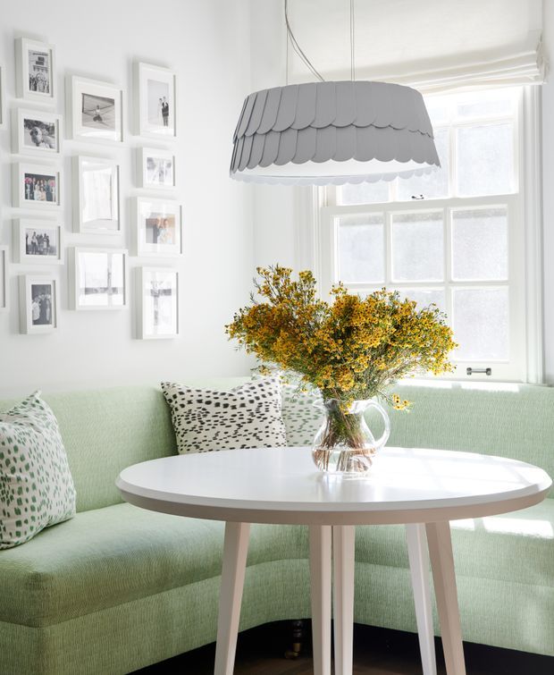 A bright, airy breakfast nook or compact dining area with a pale mint-green upholstered corner banquette, a round white table with light timber legs, and a large patterned pendant light. White-framed windows with a roller blind fill the room with natural light, while a gallery wall of framed photographs decorates the white wall. A glass vase of yellow flowers sits on the table, with patterned cushions adding visual interest.