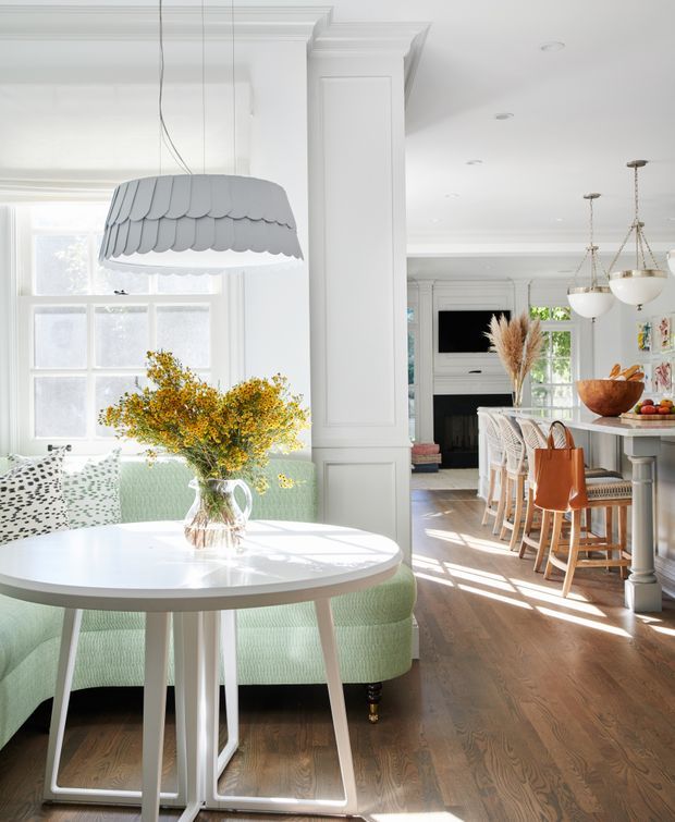 Bright, refined open-plan interior featuring a breakfast nook with a round white table, pale mint upholstered banquette, patterned cushions, and an oversized scalloped pendant light. The space opens into a kitchen and dining area with a large island, timber bar stools, white cabinetry, decorative wall panelling, classic trim, and a fireplace beyond. Warm timber flooring and abundant natural light create an airy, welcoming atmosphere.