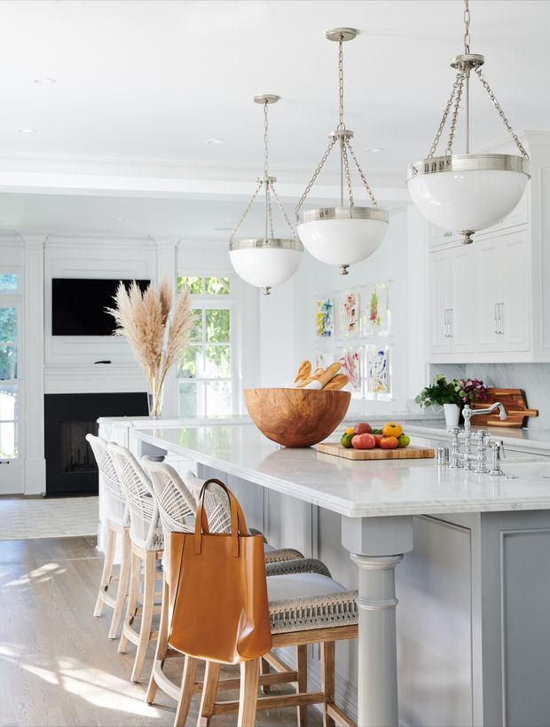 Bright, refined kitchen interior with a large white island, pale cabinetry, paneled walls, a marble-look benchtop, woven counter stools, and three hanging bowl pendant lights. A fireplace and television are visible in the adjoining living area, while large windows bring in natural light. The styling combines classic architectural detailing with warm natural timber and wicker accents.