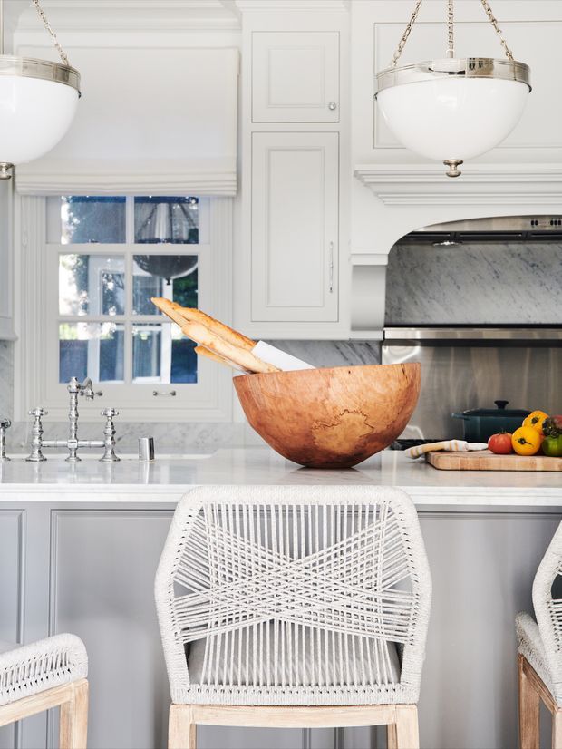 Bright, refined kitchen interior with white cabinetry, decorative panelled joinery, marble-look backsplash, pale stone island benchtop, and polished metal pendant lights. A large wooden bowl, bread, produce and woven bar stools add natural warmth to the predominantly white space. The kitchen has a classic, tailored architectural character with abundant daylight.