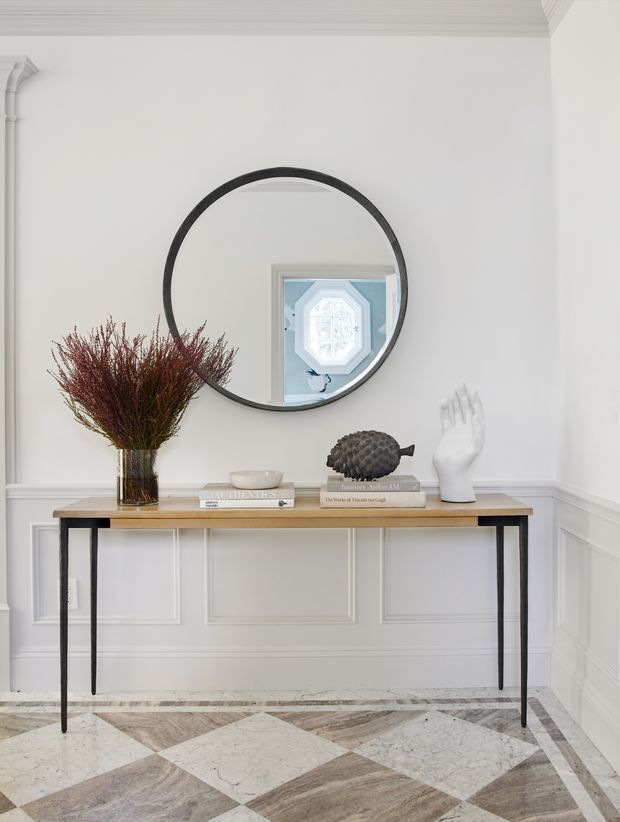 Refined entryway featuring a slim console table with a timber top and dark metal legs, positioned against white decorative wall panelling. A large round black-framed mirror hangs above the console, reflecting an octagonal window. Styling includes a vase of dried foliage, books, a small bowl, and sculptural objects. The floor has a geometric patterned stone or marble finish, and the space is bright with elegant traditional detailing.