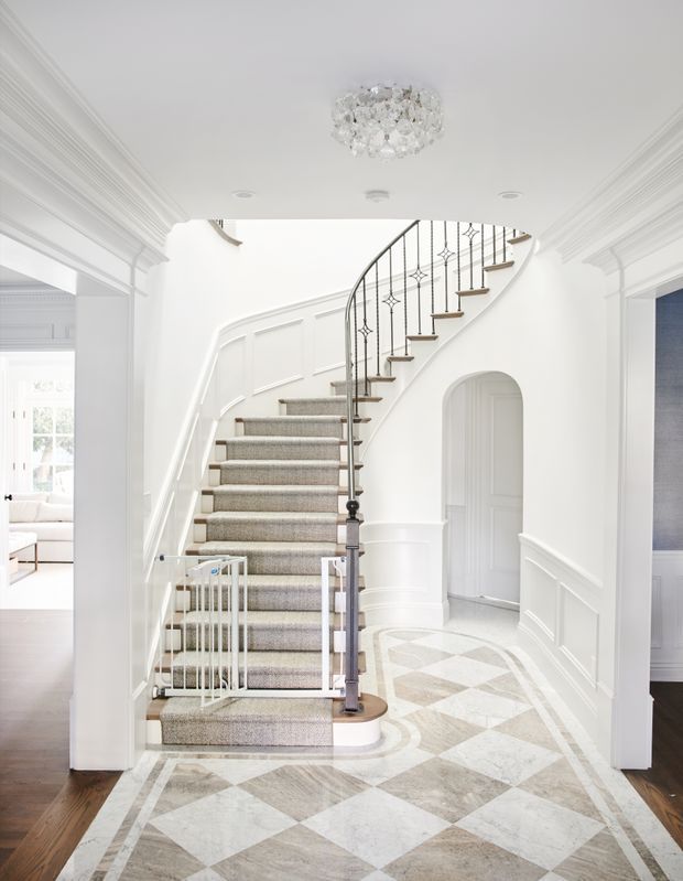 A bright, refined residential entrance hall centered on a sweeping curved staircase. The space features white panelled walls, elaborate crown moulding, decorative black metal stair balustrades, a carpeted stair runner, an arched doorway, and an ornate geometric marble or stone floor in pale grey and white. A crystal chandelier hangs from the coffered-style ceiling, with glimpses of adjoining rooms at either side.