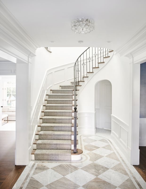Bright, refined residential entry hall centered on a curved staircase with a dark metal balustrade, timber stair treads, and a neutral carpet runner. The space features extensive white decorative wall panelling and moulded cornices, a crystal ceiling light, and an elaborate geometric stone or marble-patterned floor bordered by dark timber flooring. An arched doorway and adjoining rooms are visible.