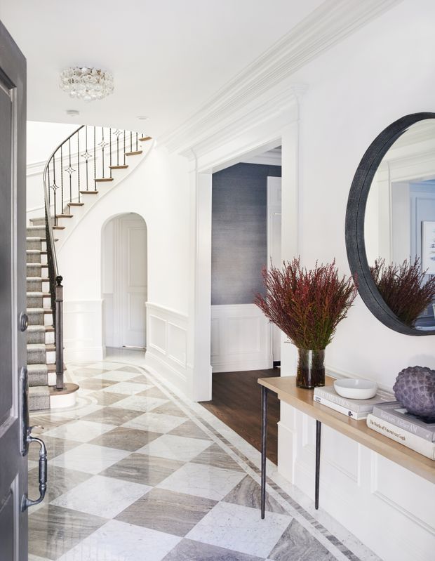 Bright, refined residential entry hall with a sweeping curved staircase, ornate white wall mouldings and wainscoting, a patterned marble or stone floor, and a slim console table beneath a large round mirror. A dark blue-grey accent wall and dark timber flooring are visible through the central doorway. The staircase has a carpet runner, dark metal balustrade, and timber handrail, while a decorative crystal ceiling light adds a traditional luxe touch.