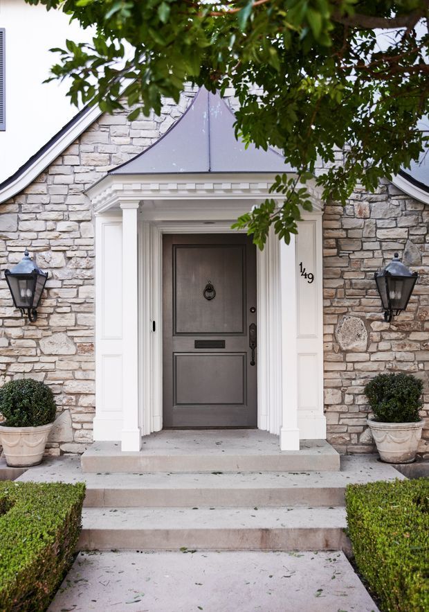 Formal residential front entry viewed from outside, featuring a dark paneled front door within an ornate white classical portico, flanked by traditional black lantern sconces. The facade is clad in irregular light-grey natural stone, with clipped hedges, symmetrical topiary planters, and concrete steps leading to the entrance. A small metal roof canopy and decorative cornice emphasize the grand, traditional approach.