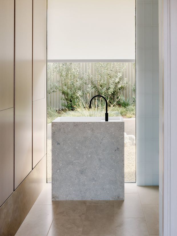 A refined contemporary kitchen centered on a substantial pale stone island with an integrated white sink and matte black gooseneck tap. Full-height light timber cabinetry lines the left wall, while pale blue-green tiled surfaces frame the right side. Large glazing or an open view behind the island looks onto a planted courtyard, and neutral large-format floor tiles create a calm, light-filled interior.