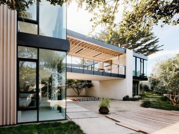 Contemporary two-storey residence with extensive black-framed glazing, pale vertical timber cladding, light masonry walls, and a projecting upper-level balcony. A timber pergola shades the open terrace, which overlooks a landscaped courtyard with gravel paving, concrete paths, planted beds, and a timber deck. The image is photographed from the exterior and emphasises the building facade and outdoor terrace.
