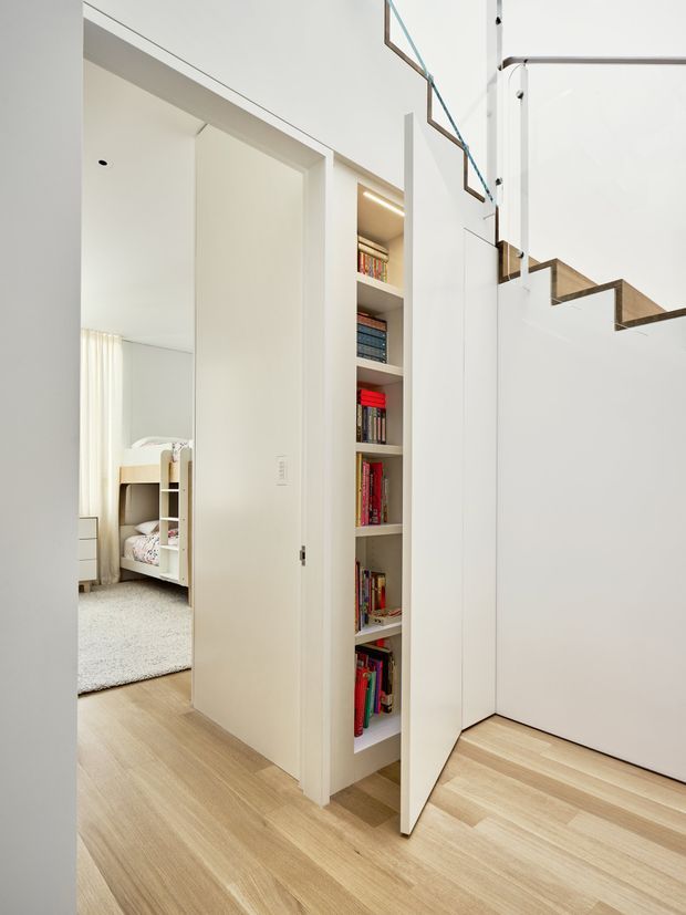 Bright contemporary interior hallway with pale timber flooring, white walls and built-in shelving filled with books. A partially open door reveals a children's bedroom, while a sculptural timber staircase with clear glass balustrade rises beside a tall white double-height wall.
